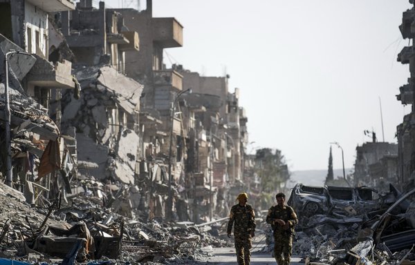 Fighters of the Syrian Democratic Forces (SDF) walk down a street in al-Raqa past destroyed vehicles and heavily damaged buildings on October 20, 2017, after a Kurdish-led force expelled ISIS from the northern Syrian city, formerly their 'capital'. [Bulent Kilic/AFP]