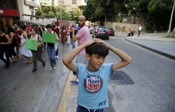 A Syrian refugee child, who works as a street vendor, looks at protesters holding banners on July 18th, 2016 in Beirut during a demonstration to protest against racism and in particular against a curfew that prevents Syrian refugees from leaving their homes between sunset and sunrise. [Joseph Eid/AFP]