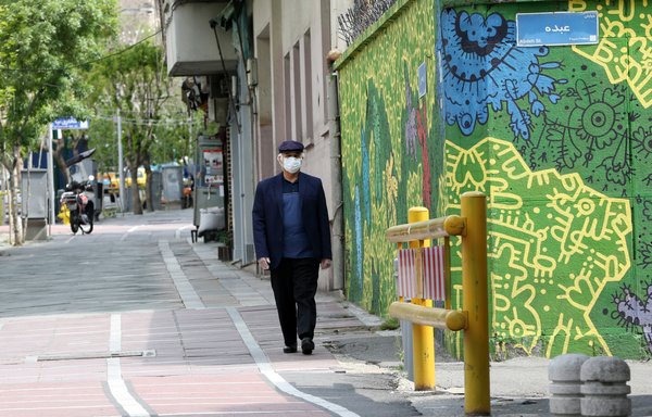 رجل إيراني يضع قناعا واقيا وهو يسير أمام رسم جداري في شارع جانبي بالعاصمة طهران يوم 13 أبريل/نيسان في قمة تفشي جائحة فيروس كورونا المستجد. [عطا كيناري/وكالة الصحافة الفرنسية]